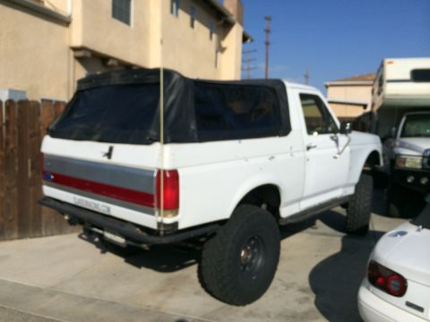 1990 Ford Bronco - Rear quarter view of a white lifted Ford SUV with black soft top and red lower tailgate trim parked between a fence and an RV.