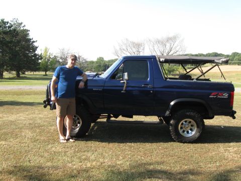 1982 Ford Bronco - A blue 1982 Ford Bronco is shown from the side with a man standing next to it in a grassy field.