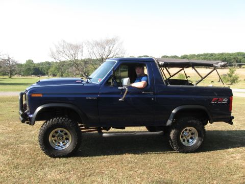 1982 Ford Bronco - A man sits in a dark blue Ford Bronco with a soft top and off-road tires.