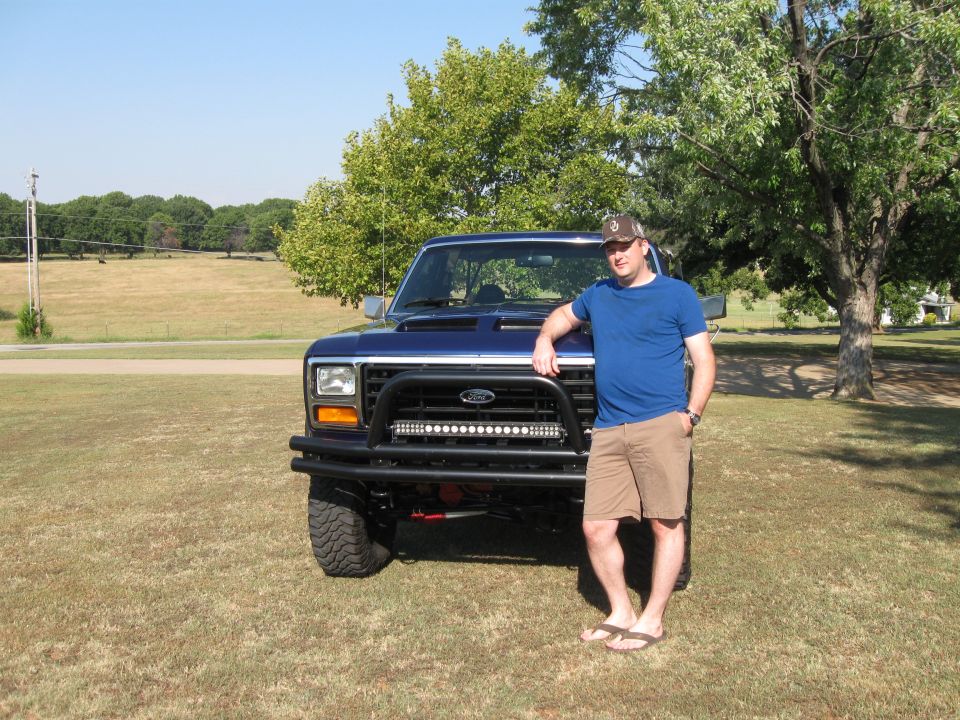 Man leaning on blue Ford truck with aftermarket bumper and light bar. Truck has square headlights and body lines consistent with early 1980s Ford