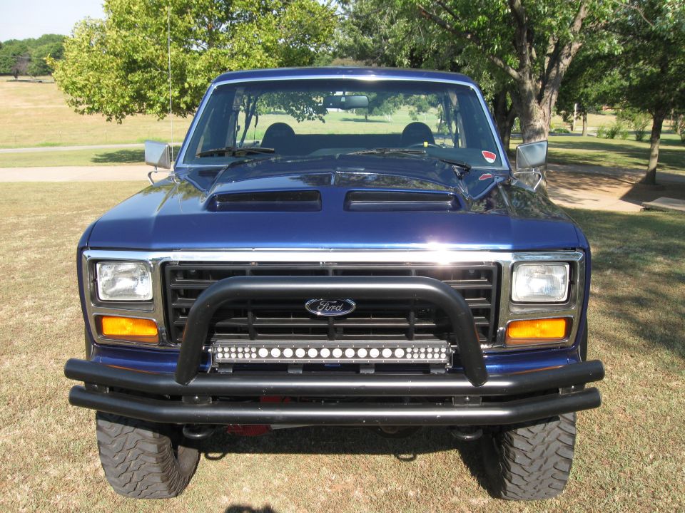 Front view of a blue Ford Bronco with a custom hood scoop, bull bar, and LED light bar. It has a large aftermarket bumper and off-road tires