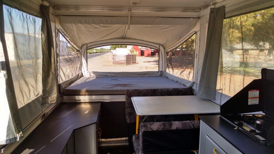 Interior of a pop-up camper with a bed, table, and stove. Curtains are drawn on the sides