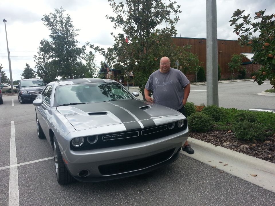Man standing next to a silver 2015 Dodge Challenger with black racing stripes on the hood and roof