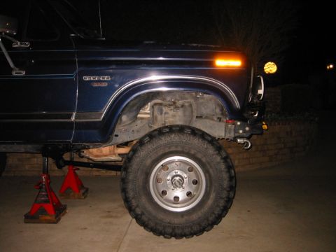 1985 Ford Bronco - The front quarter panel of a dark blue Ford Bronco is shown, with the front wheel and suspension components visible.