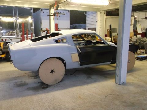 1967 Ford Mustang - A primer-gray 1967 Ford Mustang body sits in a garage, awaiting restoration, with its driver's door ajar and wooden wheels in place.