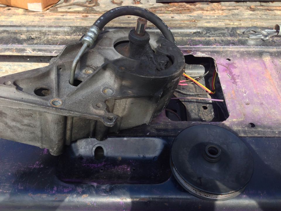 Power steering pump assembly with hose and pulley removed, sitting on a vehicle's chassis