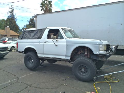 1990 Ford Bronco - white lifted Ford Bronco-style SUV sits in a paved lot with its front wheel removed and suspension exposed.