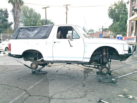 1990 Ford Bronco - white SUV with soft top is raised on jacks with both wheels off, exposing the coil-spring front suspension in a lot.