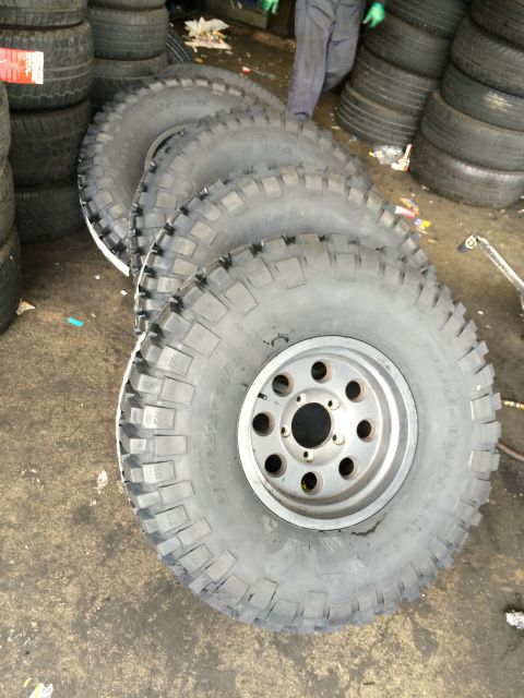 1990 Ford Bronco - Four chunky off-road tires lean on a shop floor, the front one mounted on a silver eight-lug wheel.