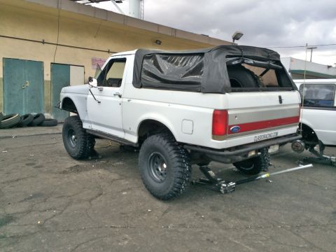 1990 Ford Bronco - Rear three-quarter view of a white lifted Ford SUV with black soft top and red trim parked in an industrial yard under grey skies.