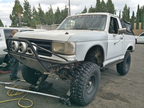1990 Ford Bronco - Front three-quarter of a white lifted Ford SUV with a tube prerunner bumper and round driving lights, front wheel removed in a lot.