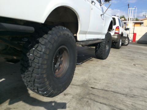 1990 Ford Bronco - Low-angle view of a mud tire on a bronze wheel under a white lifted truck, with an SUV parked nearby in a lot.