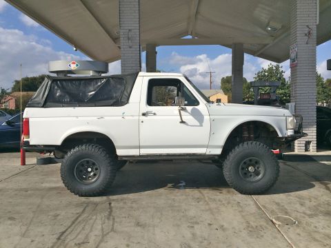 1990 Ford Bronco - Side profile of a white lifted Ford SUV with black soft top parked under a gas-station canopy on a sunny day.