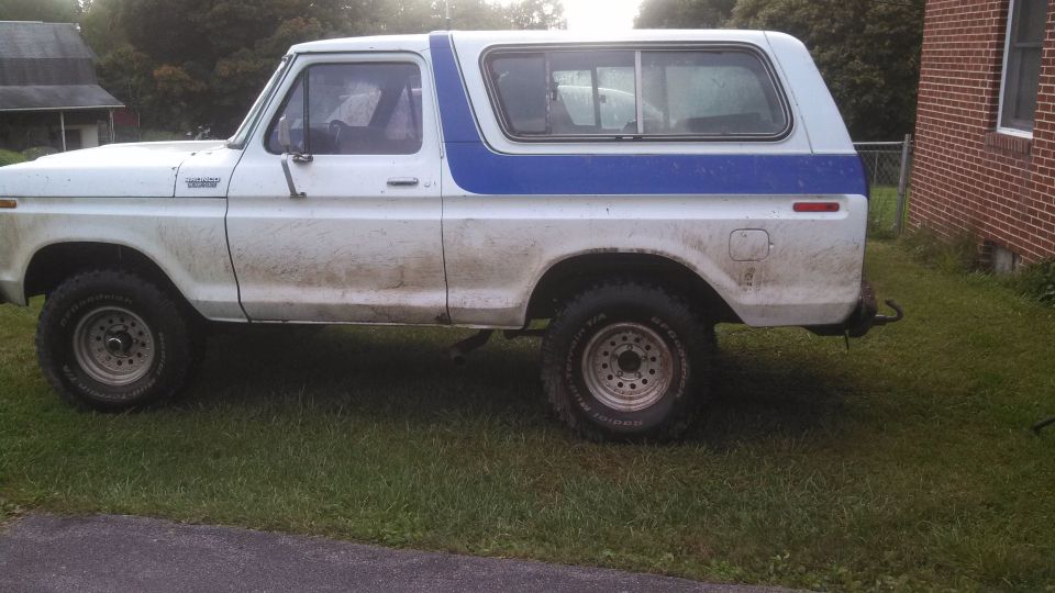 Side view of a white and blue Ford Bronco parked on grass. The vehicle has mud splatters on its body