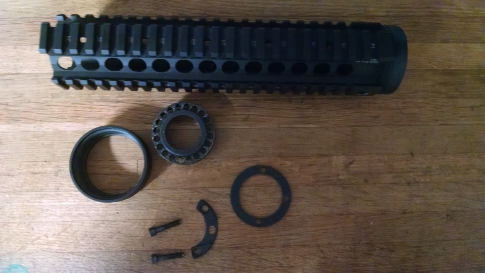 Black gun barrel shroud with accessory rail, a ring, a toothed ring, two screws, and two curved metal pieces on a wooden surface