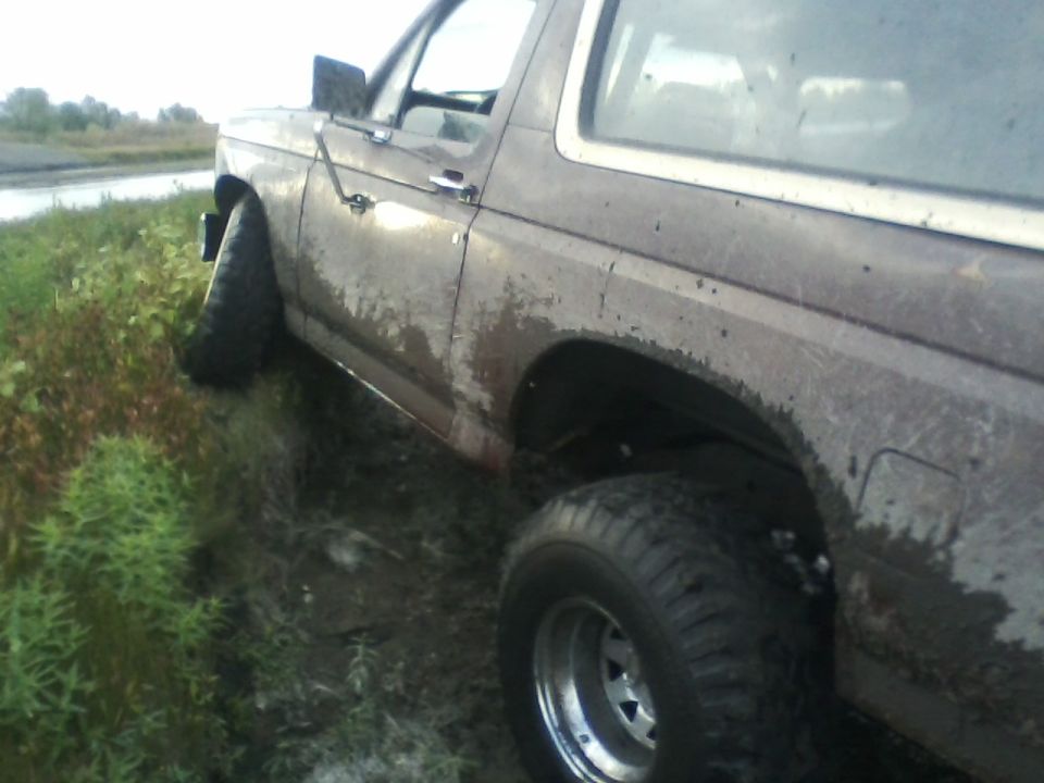 Brown 1980 Ford Bronco off-road, mud-splattered, with a white roof trim and chrome wheels