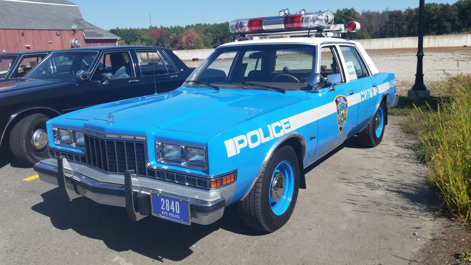 Blue and white police car with "POLICE" text and "NYC POLICE" license plate, parked next to a black car