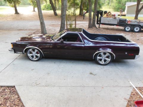 1979 Ford Ranchero - Driver-side profile of a lowered dark burgundy/plum coupe-utility parked on a concrete driveway under oak trees, wearing large chrome multi…