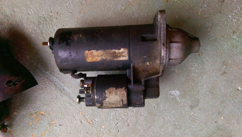 Black starter motor with two labels, one partially obscured, resting on a concrete surface