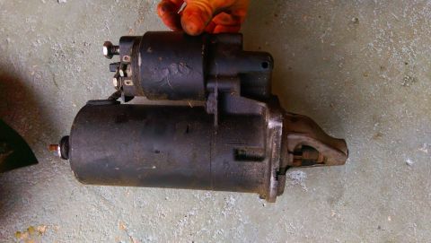 1989 BMW 3 Series - A black starter motor is shown resting on a concrete surface, held by a hand at the top.