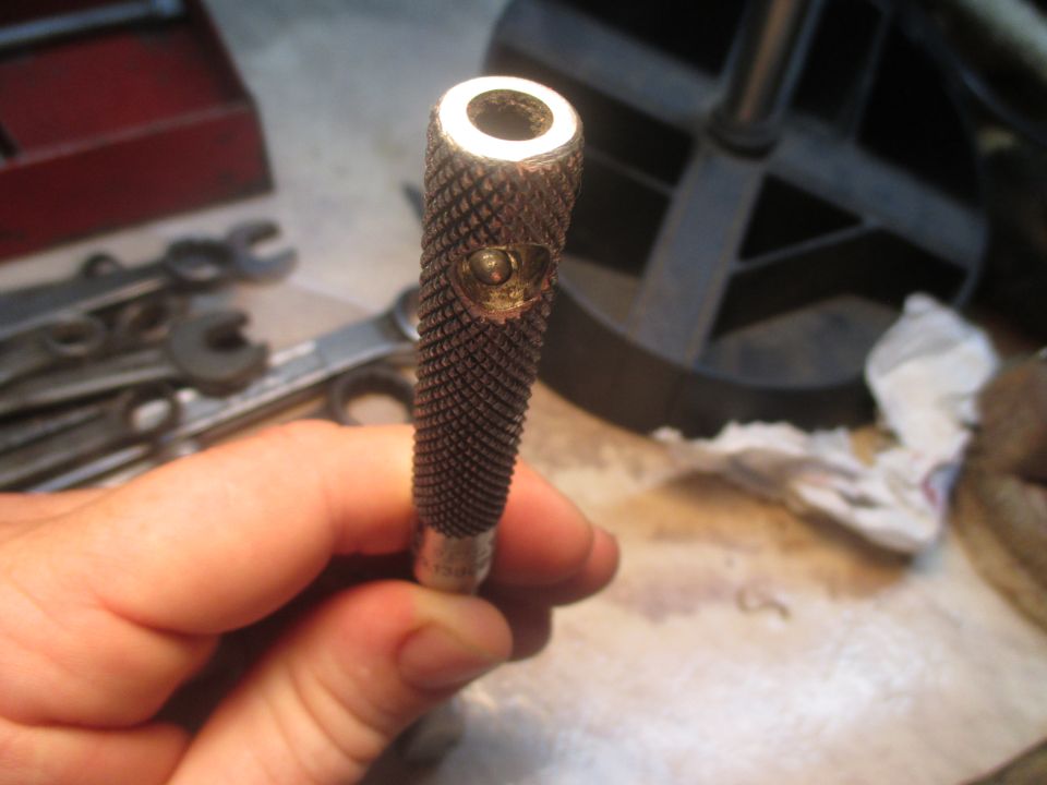 Knurled metal tool part held in hand, with a circular opening at the top and a small circular indentation below it