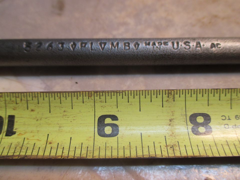 Close-up of a metal rod with stamped text "8263 PLVMBO MADE IN U.S.A. AC" next to a yellow measuring tape showing inches