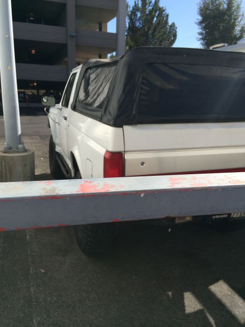 1990 Ford Bronco - Rear view of a white lifted Ford SUV with black soft top, partly framed by a guardrail, beside a concrete parking structure.