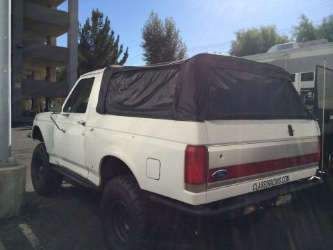 1990 Ford Bronco - Rear quarter view of a white lifted Ford SUV with black soft top and CLASS8RACING.COM plate frame near a parking structure.