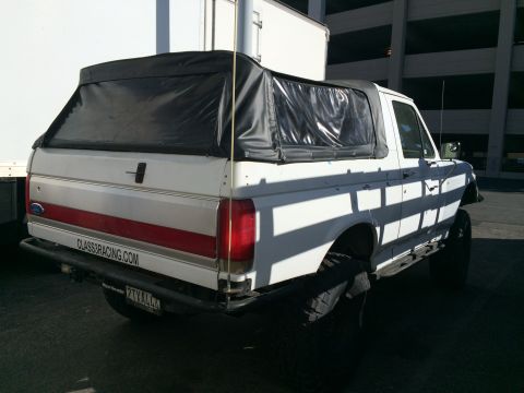 1990 Ford Bronco - Rear three-quarter view of a white lifted Ford SUV with black soft top, red trim and a CLASS8RACING.COM plate frame in a paved lot.