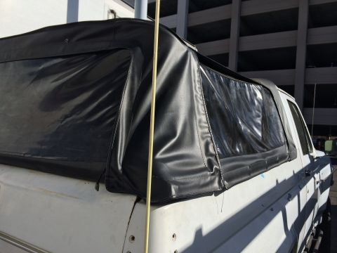 1990 Ford Bronco - Close-up of a black vinyl soft top with zippered plastic windows fitted over the rear of a white SUV in sunlight.