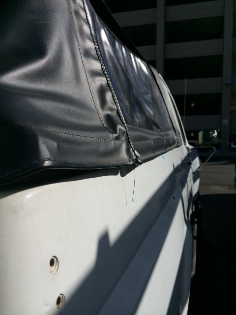 1990 Ford Bronco - Close-up of the stitched corner of a black vinyl soft top meeting the white painted body panel of a vehicle in sunlight.