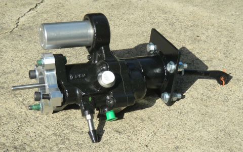 1982 Ford Bronco - This black power steering gearbox features a silver cylinder and is mounted on a metal bracket, shown against a concrete surface.