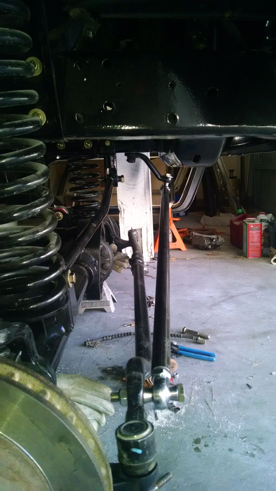 Close-up of vehicle suspension components, including springs and steering linkage, in a garage setting. Watermark reads SUPERMOTORS.NET