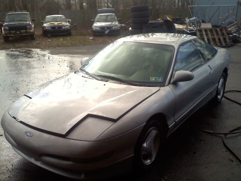 1993 Ford Probe - A silver 1993 Ford Probe GT is parked on a wet surface, with other vehicles and stacked tires visible in the background.