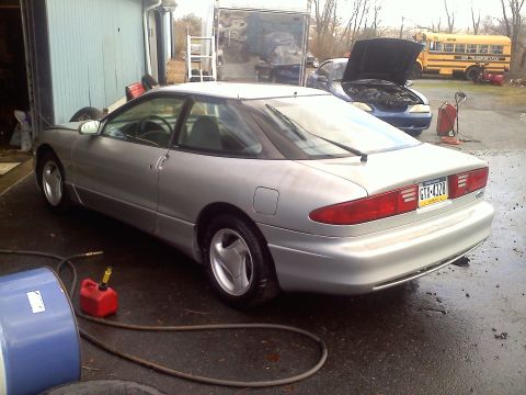 1993 Ford Probe - A silver 1993 Ford Probe GT is parked outside a garage, with another vehicle and a school bus in the background.