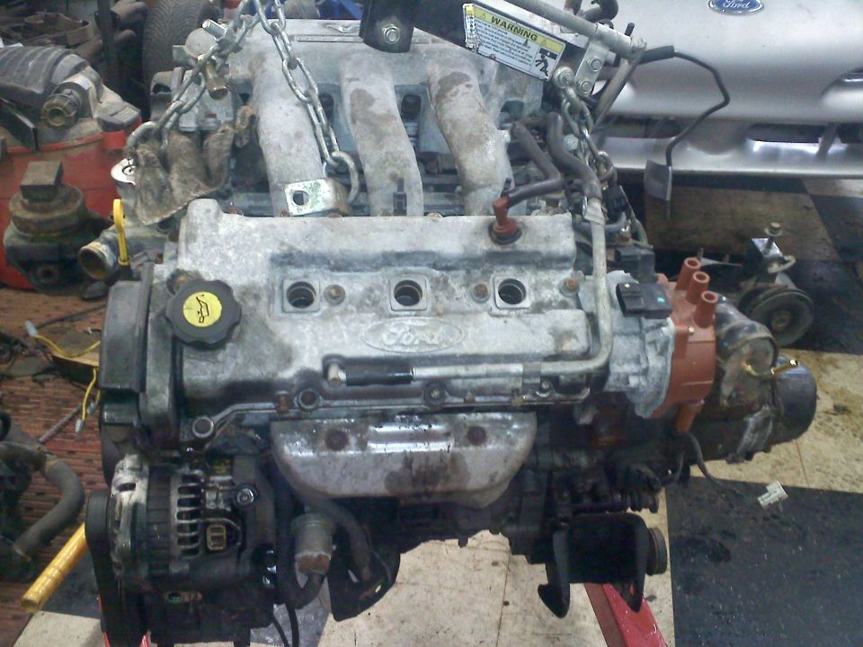 Ford engine assembly, possibly from a car, suspended by chains. It shows the intake manifold, valve cover with Ford logo, alternator, and distributor