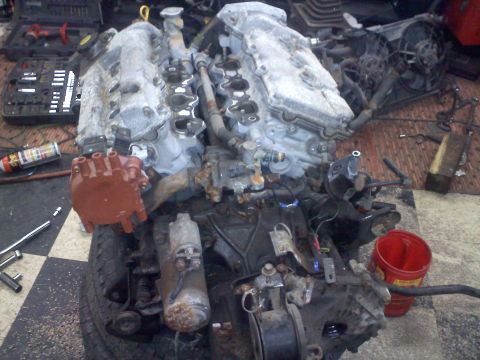 1993 Ford Probe - A V6 engine is disassembled on a checkered floor, with tools and a red bucket nearby.
