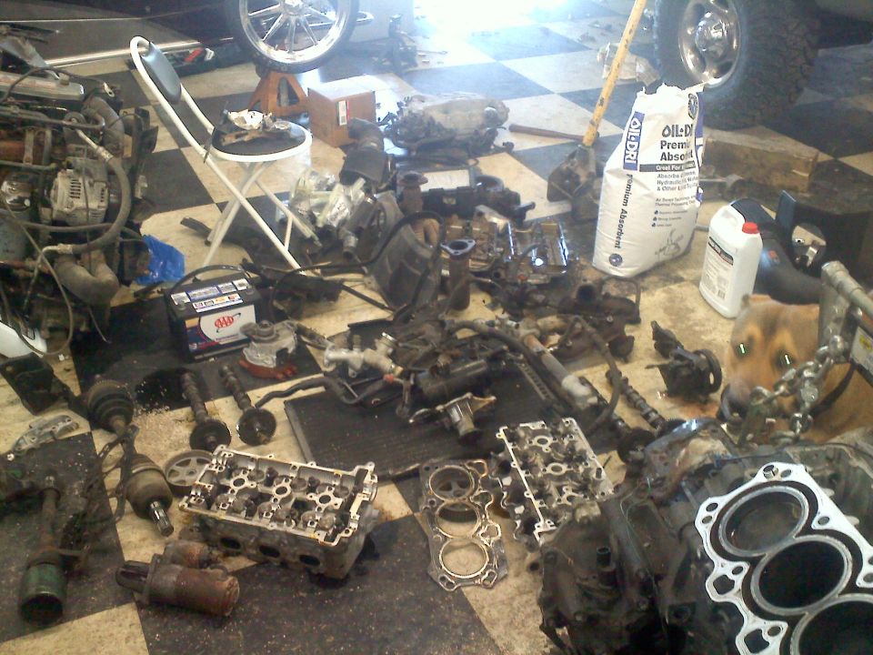 Disassembled engine parts and a car battery on a checkered floor, with a dog in the background