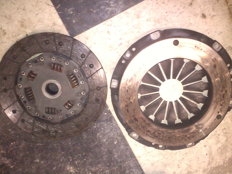 Clutch disc and pressure plate on checkered floor. The disc has springs and a splined center. The pressure plate has radiating fins