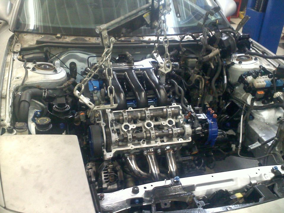 Engine being lifted out of a car with chains and an engine hoist. The engine has exposed camshafts and exhaust headers
