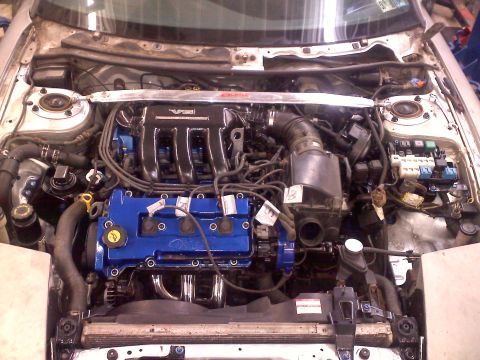 1993 Ford Probe - The engine bay of a car is shown, a V6 engine with blue painted valve covers and chrome exhaust headers.