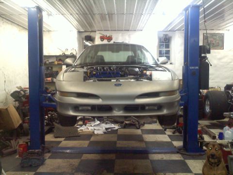 1993 Ford Probe - A silver 1993 Ford Probe GT is raised on a two-post lift in a cluttered garage, its hood open revealing the engine.