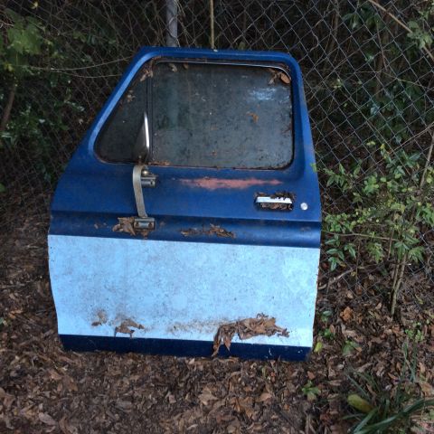 1979 Ford Bronco - A single weathered Ford truck door is shown, detached from a vehicle and resting on the ground.
