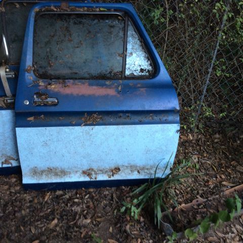 1979 Ford Bronco - Two weathered truck doors, possibly from an older model, lie discarded outdoors near a fence.
