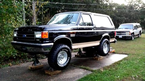 1991 Ford Bronco - A black 1991 Ford Bronco is elevated on jack stands, with another SUV parked behind it in a driveway.