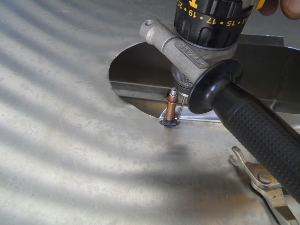 Drill bit entering a circular hole in a brushed metal panel, with a wrench visible below