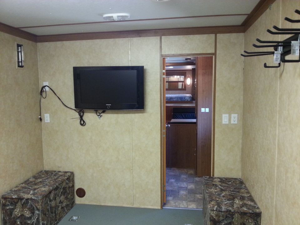 Interior of a trailer showing a mounted TV, camouflage storage benches, and a doorway leading to bunk beds