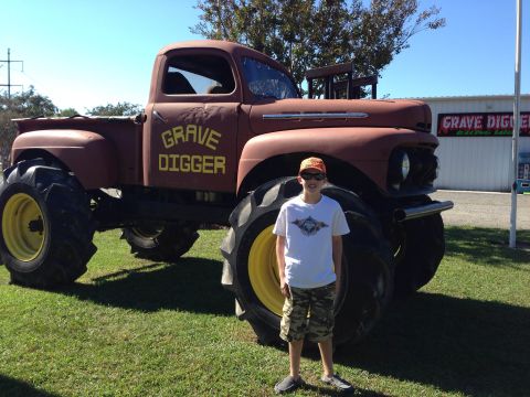 1979 Ford Bronco - A boy stands next to a custom monster truck painted with "Grave Digger" on its side, massive tires.