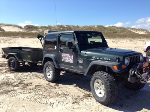 1979 Ford Bronco - A dark green Jeep Wrangler Rubicon is parked on a sandy beach, towing a utility trailer.