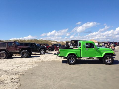 1979 Ford Bronco - Several Jeeps, including a green pickup, are parked on a sandy beach under a blue sky.
